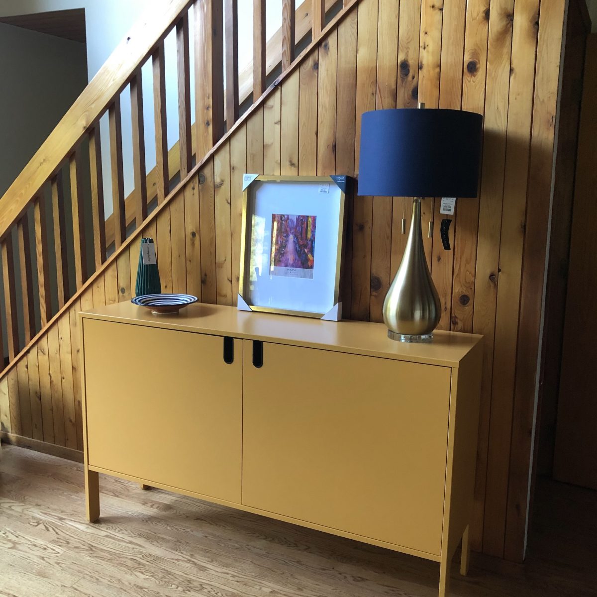 A different photo of the original cabinet, but with actual decorative items on its surface. (Not ai-generated.) On the surface is a decorative green vase, a decorative blue bowl, a medium-sized picture with a gold frame, and a gold teardrop-shaped lamp with a dark blue shade.